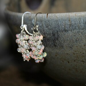 Peut inclure: Une paire de boucles d'oreilles pendantes en argent avec un groupe de perles d'opale blanche iris&eacute;e. Les boucles d'oreilles pendent d'un bol en c&eacute;ramique gris.