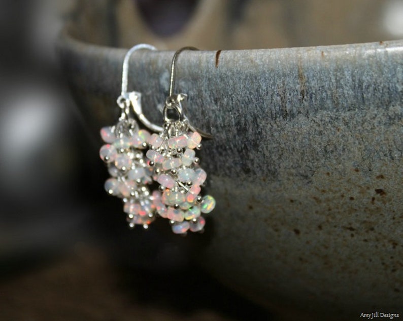 Peut inclure: Une paire de boucles d'oreilles pendantes en argent avec un groupe de perles d'opale blanche iris&eacute;e. Les boucles d'oreilles pendent d'un bol en c&eacute;ramique gris.
