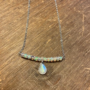 May include: A delicate silver necklace featuring a bar of small, iridescent beads and a teardrop-shaped pendant. The pendant and beads have a rainbow sheen. The necklace is displayed on a wooden surface.