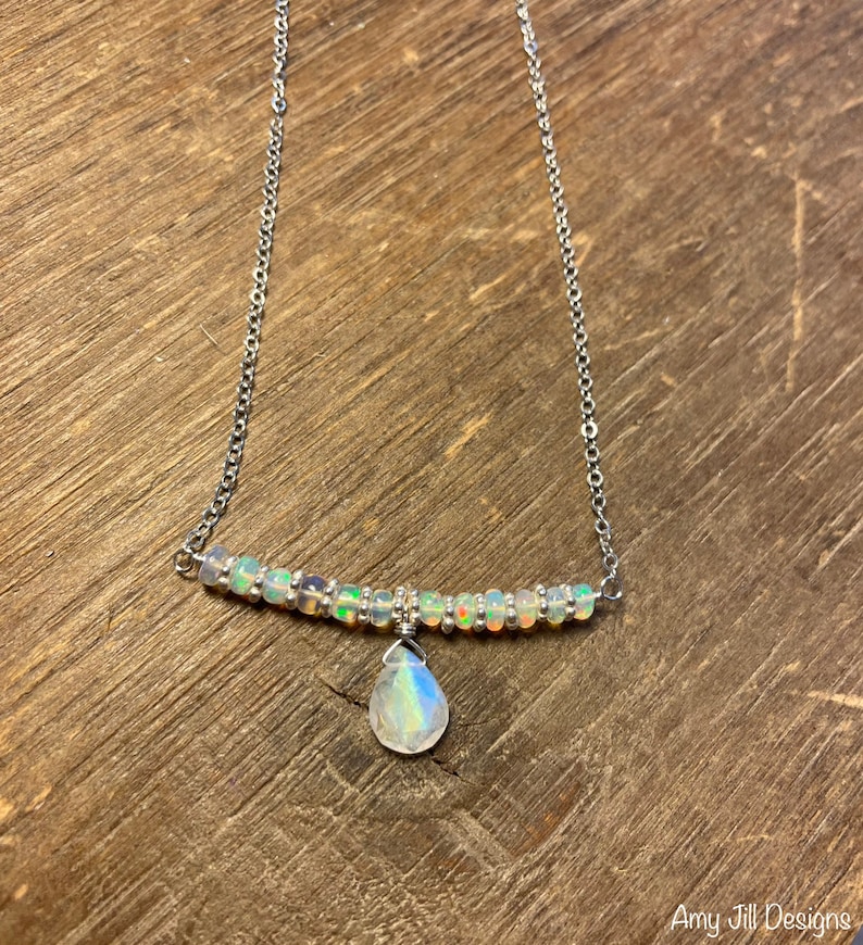 May include: A delicate silver necklace featuring a bar of small, iridescent beads and a teardrop-shaped pendant. The pendant and beads have a rainbow sheen. The necklace is displayed on a wooden surface.