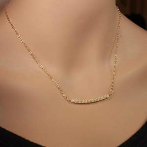 May include: A gold chain necklace with a delicate row of small, clear stones forming a curved bar pendant.