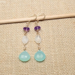 May include: A pair of dangle earrings featuring a gold-colored wire design. Each earring has a purple bead, a white bead, and a light blue teardrop-shaped bead. The earrings are displayed on a light brown surface.