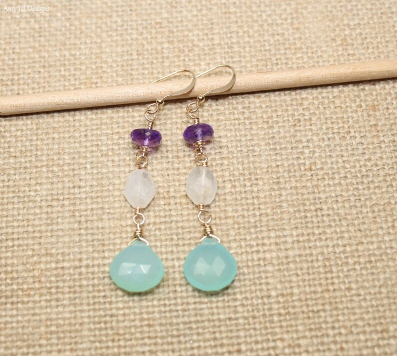 May include: A pair of dangle earrings featuring a gold-colored wire design. Each earring has a purple bead, a white bead, and a light blue teardrop-shaped bead. The earrings are displayed on a light brown surface.