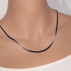 May include: A delicate necklace featuring a gradient of black, blue, and light blue beads. The necklace is displayed on a person wearing a white tank top. The beads appear to be small and evenly spaced, creating a subtle, elegant design.