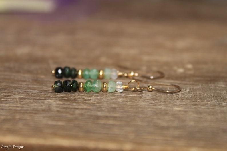 May include: A pair of dangle earrings featuring a gradient of green gemstone beads and gold spacers. The earrings have gold hooks.