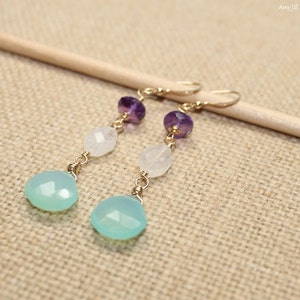 May include: A pair of dangle earrings featuring a gold-tone hook, amethyst, moonstone, and teardrop-shaped aqua chalcedony beads. The earrings are displayed on a neutral-toned surface.