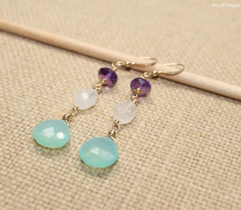 May include: A pair of dangle earrings featuring a gold-tone hook, amethyst, moonstone, and teardrop-shaped aqua chalcedony beads. The earrings are displayed on a neutral-toned surface.