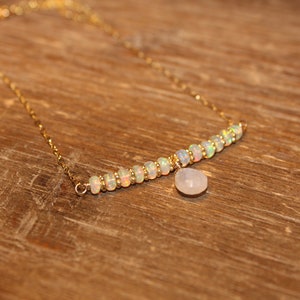 May include: A gold necklace with a row of opal beads and a small, pear-shaped, white gemstone pendant. The necklace is presented on a wooden surface, highlighting its elegant design and craftsmanship.