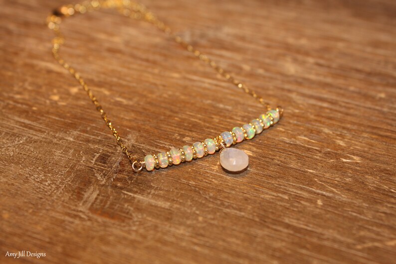 May include: A gold necklace with a row of opal beads and a small, pear-shaped, white gemstone pendant. The necklace is presented on a wooden surface, highlighting its elegant design and craftsmanship.