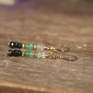 May include: A pair of green and black gemstone earrings with gold accents. The earrings are made with a variety of green and black gemstones, including emerald, peridot, and onyx. The earrings are finished with gold-plated hooks.