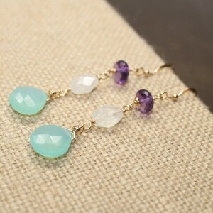 May include: A pair of dangle earrings featuring a light blue teardrop-shaped gemstone, white gemstone, and purple amethyst beads. The gemstones are strung on gold-colored wire with a hook closure. The earrings are displayed on a tan fabric surface.