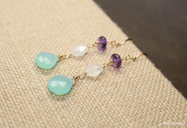 May include: A pair of dangle earrings featuring a light blue teardrop-shaped gemstone, white gemstone, and purple amethyst beads. The gemstones are strung on gold-colored wire with a hook closure. The earrings are displayed on a tan fabric surface.
