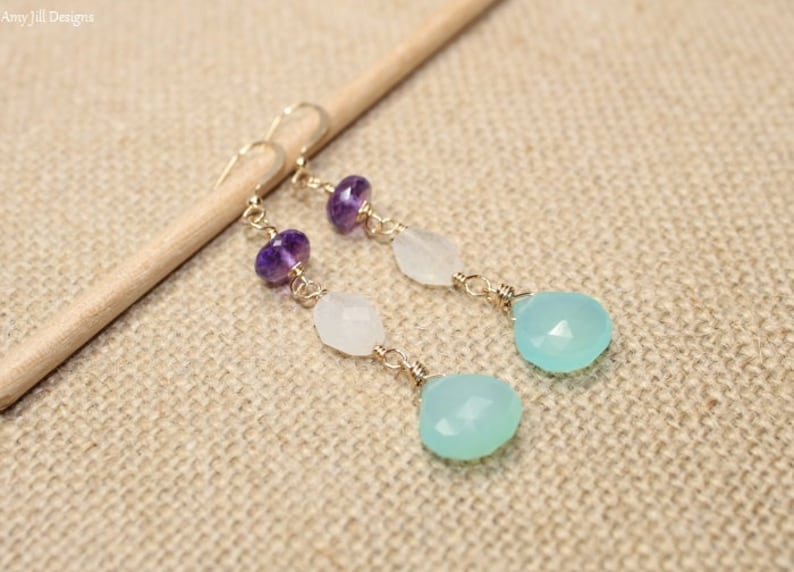 May include: Dangling earrings with gold-colored wire and a variety of gemstones. The earrings feature amethyst, white, and light blue teardrop-shaped stones. The earrings are displayed on a wooden dowel against a neutral background.