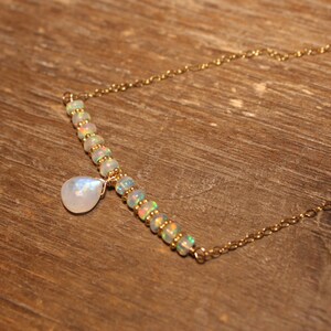 May include: A gold necklace with a bar of opal beads and a moonstone pendant. The necklace is displayed on a wooden surface. The opal beads have a rainbow sheen, and the moonstone is teardrop-shaped. The necklace is a beautiful piece of jewelry.