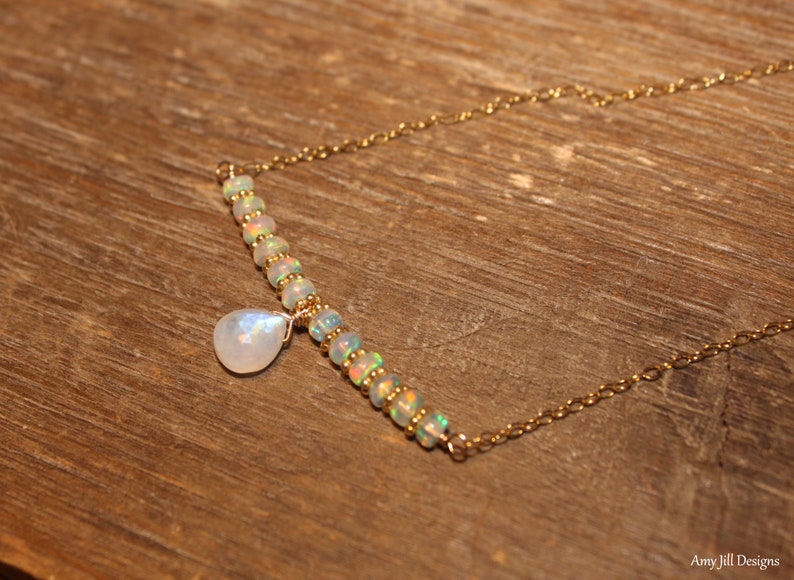 May include: A gold necklace with a bar of opal beads and a moonstone pendant. The necklace is displayed on a wooden surface. The opal beads have a rainbow sheen, and the moonstone is teardrop-shaped. The necklace is a beautiful piece of jewelry.