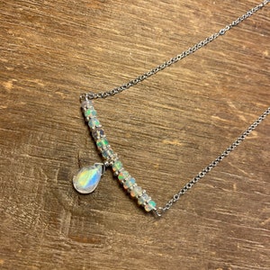 May include: A silver necklace with a bar of small, iridescent beads and a teardrop-shaped pendant. The necklace is presented on a wooden surface, highlighting its delicate sparkle and elegant design.