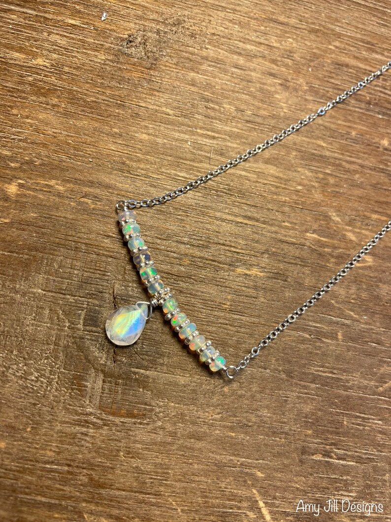 May include: A silver necklace with a bar of small, iridescent beads and a teardrop-shaped pendant. The necklace is presented on a wooden surface, highlighting its delicate sparkle and elegant design.