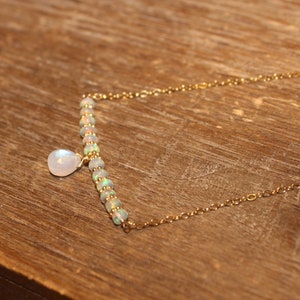 May include: A delicate gold necklace featuring a bar of small, iridescent opal beads and a teardrop-shaped moonstone pendant. The necklace is on a gold chain, and the design is simple and elegant. The background is a wooden surface.