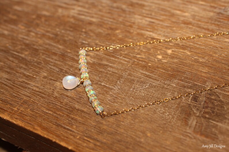 May include: A delicate gold necklace featuring a bar of small, iridescent opal beads and a teardrop-shaped moonstone pendant. The necklace is on a gold chain, and the design is simple and elegant. The background is a wooden surface.