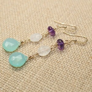 May include: Dangle earrings featuring a teardrop-shaped, light blue gemstone, white gemstone, and purple amethyst beads. The earrings are connected with gold-colored wire and have a hook closure. The earrings are displayed on a tan fabric surface.