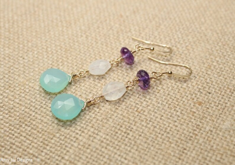 May include: Dangle earrings featuring a teardrop-shaped, light blue gemstone, white gemstone, and purple amethyst beads. The earrings are connected with gold-colored wire and have a hook closure. The earrings are displayed on a tan fabric surface.