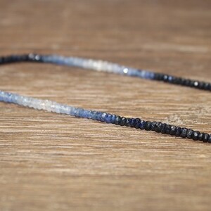 May include: A delicate necklace composed of small, faceted beads in a gradient of blue hues, transitioning from dark navy to light blue and white. The necklace is displayed on a wooden surface, showcasing its color variations.
