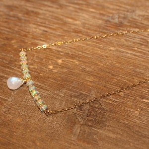 May include: A delicate gold necklace with a bar of opal beads and a pear-shaped moonstone pendant. The necklace is on a wooden surface. The chain is approximately 45 cm long.