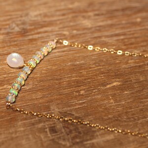 May include: A gold necklace with a bar of opal beads and a moonstone pendant. The necklace has a fine gold chain. The jewellery is set against a wooden background. The necklace is elegant and suitable for everyday wear.