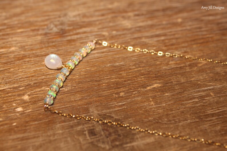 May include: A gold necklace with a bar of opal beads and a moonstone pendant. The necklace has a fine gold chain. The jewellery is set against a wooden background. The necklace is elegant and suitable for everyday wear.