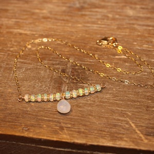 May include: A gold necklace featuring a bar of opal beads and a small, pear-shaped pendant. The necklace is presented on a wooden surface. The gold chain and opal beads create a subtle, elegant appearance.