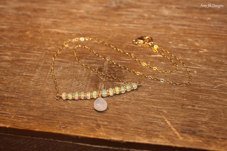 May include: A gold necklace featuring a bar of opal beads and a small, pear-shaped pendant. The necklace is presented on a wooden surface. The gold chain and opal beads create a subtle, elegant appearance.