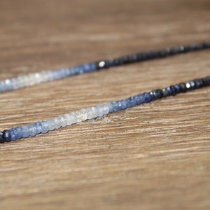 May include: A delicate necklace featuring a gradient of blue sapphire beads. The beads transition from dark navy to light blue and white, creating a subtle ombre effect. The necklace is displayed on a wooden surface.
