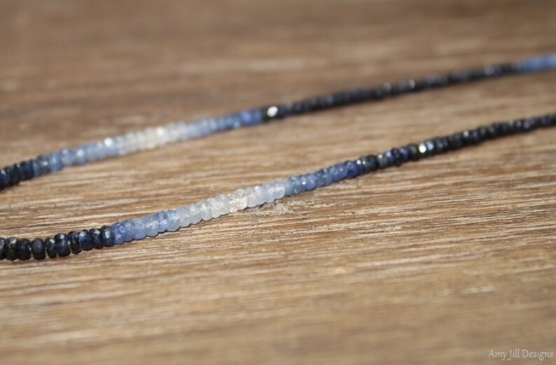 May include: A delicate necklace featuring a gradient of blue sapphire beads. The beads transition from dark navy to light blue and white, creating a subtle ombre effect. The necklace is displayed on a wooden surface.