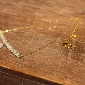 May include: A delicate gold necklace with a bar pendant featuring small, iridescent beads and a teardrop-shaped charm. The necklace is displayed on a wooden surface, showcasing its elegant design.