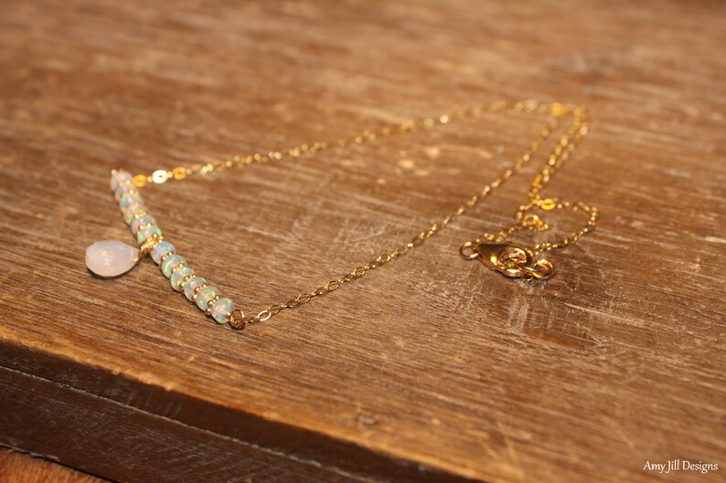 May include: A delicate gold necklace with a bar pendant featuring small, iridescent beads and a teardrop-shaped charm. The necklace is displayed on a wooden surface, showcasing its elegant design.