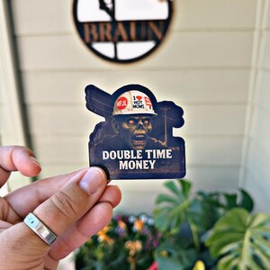 May include: A sticker featuring a zombie construction worker wearing a hard hat with the text "I ❤️ HOT MOMS" and "MFJL". The sticker also has the words "DOUBLE TIME MONEY". The background includes a house number sign and potted plants.