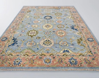 May include: A rectangular area rug with a light blue field and a detailed floral pattern in shades of pink, green, and blue. The rug has a border in a complementary color palette. The rug is made of a soft material.