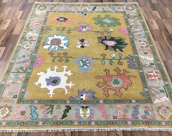 May include: Mustard yellow area rug with a central design featuring various floral and geometric shapes in white, pink, green, and orange. The rug has a border with a repeating pattern in green and tan.