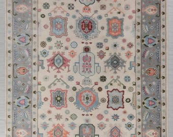 May include: Rectangular area rug with a cream-colored field and a gray border. The rug features a repeating pattern of geometric shapes in shades of pink, blue, orange, and gray. The design includes floral and abstract motifs.