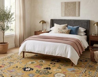 May include: A bedroom featuring a dark gray upholstered bed frame with a white duvet and a dusty rose waffle-knit throw blanket. The bed is styled with cream and blue pillows. A large mustard yellow area rug with a floral pattern covers the floor.