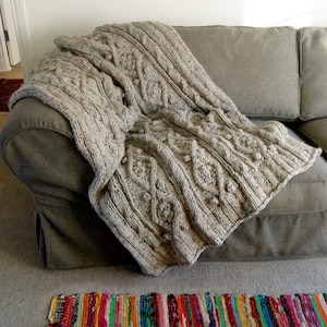 May include: A large, knitted throw blanket in a light brown color with a cable knit pattern. The blanket is draped over a gray couch.
