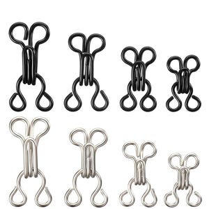 Op de afbeelding: Een assortiment zwarte en zilveren metalen haak- en oogsluitingen. De sluitingen zijn in verschillende maten, met een lusvormige bovenkant en een gebogen onderkant. De haken zijn ontworpen voor naaien en kledingconstructie.