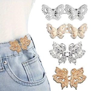 May include: A set of four metal butterfly-shaped waist cinchers in silver and gold tones, featuring intricate cut-out wing designs and embedded clear rhinestones. One cincher is shown attached to the waistband of light blue denim jeans.