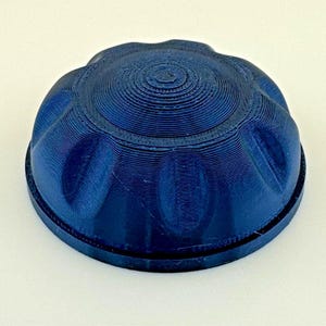 May include: A dark blue, circular, dome-shaped cap with a ribbed, scalloped edge and a textured, concentric ring pattern on the top surface. The object features a 3D-printed finish with visible layer lines throughout its entire structure.