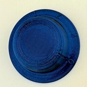 May include: A dark blue circular object with a textured surface featuring concentric rings and raised numbers 1, 2, and 3 along the outer edge. The center has a diagonal line pattern, and a small loop is attached to the side of the rim.
