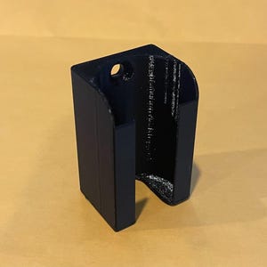 May include: A black 3D printed wall mount bracket with a U-shaped open front design and a single circular mounting hole at the top. The object features a flat back panel and vertical side walls with a textured finish from the printing process.