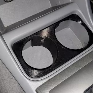 May include: A black, dual-cup holder insert designed for a vehicle center console. The accessory features two circular openings with a smooth, matte finish, specifically shaped to fit into the existing gray plastic console compartment.