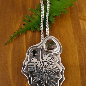 Pu&ograve; includere: Una collana d'argento con un ciondolo che presenta un designo a foglia dettagliato, accentuato da una pietra preziosa verde e marrone e una pietra preziosa nera.
