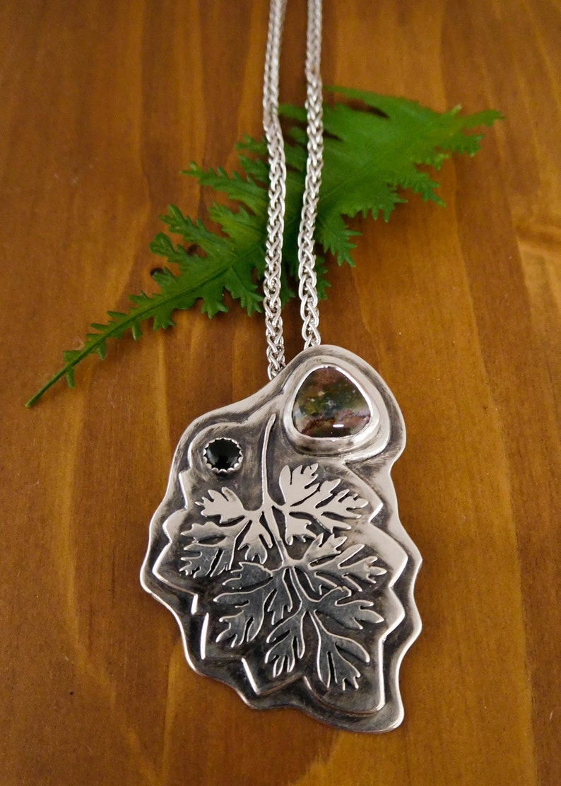 Pu&ograve; includere: Una collana d'argento con un ciondolo che presenta un designo a foglia dettagliato, accentuato da una pietra preziosa verde e marrone e una pietra preziosa nera.