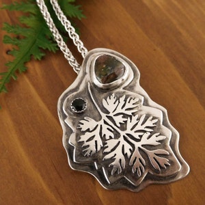 Pu&ograve; includere: Una collana con un ciondolo in argento che presenta un designo dettagliato di foglia di felce con una pietra verde e marrone e una pietra nera.